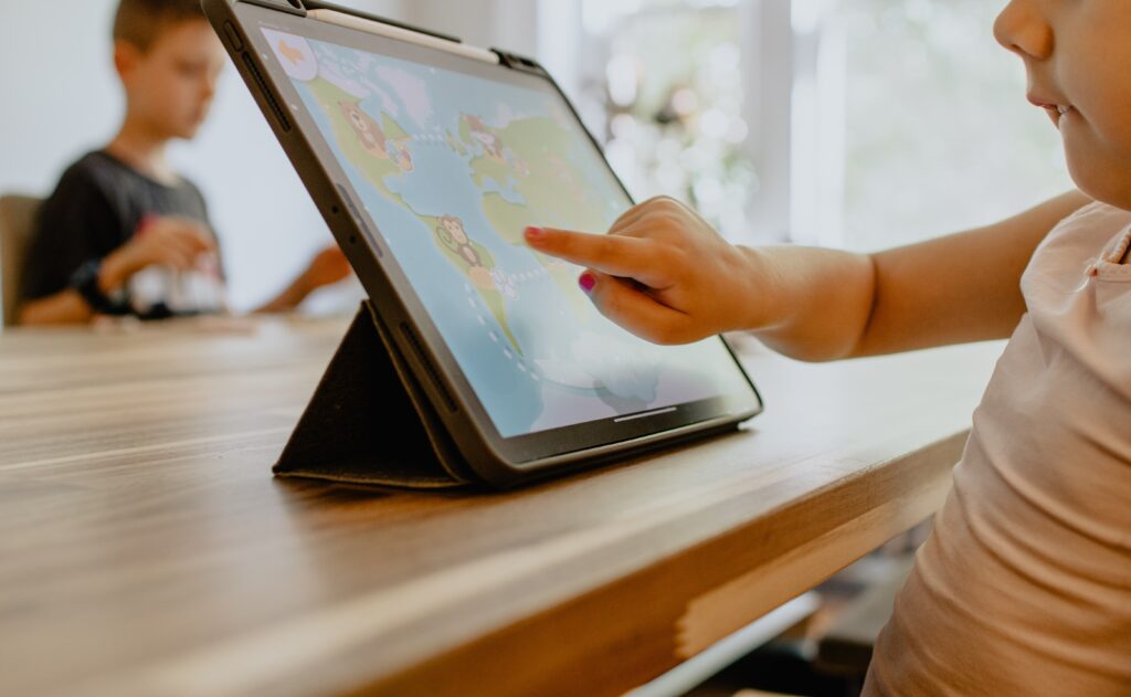 Two kids of different ages at a table, one on a tablet interacting with a map.